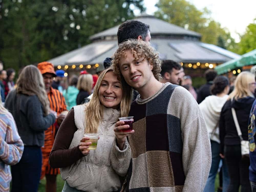 Two people stand close together, smiling and holding drinks at an outdoor gathering during Woodland Park Zoo’s WildLanterns Night Owl nights. A crowd and the iconic round building can be seen in the background, capturing the lively atmosphere of one of Seattle’s premier 21+ events.