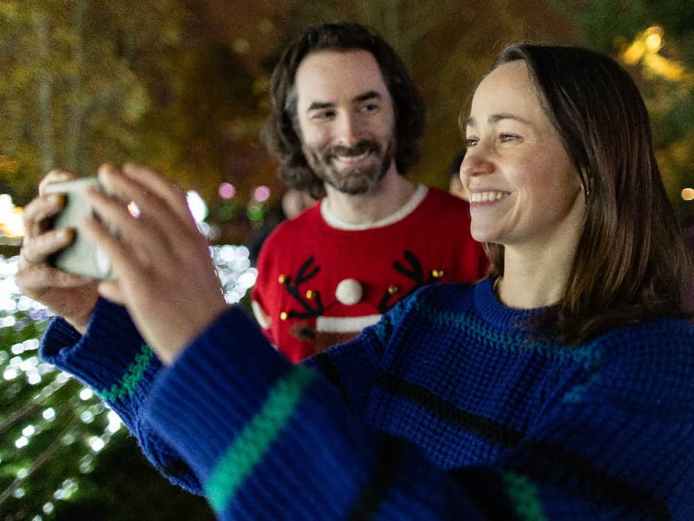 A woman takes a selfie with a man in a festive sweater outdoors at night, surrounded by holiday lights at Woodland Park Zoo during OktoBEARfest, one of Seattle’s favorite 21+ events.