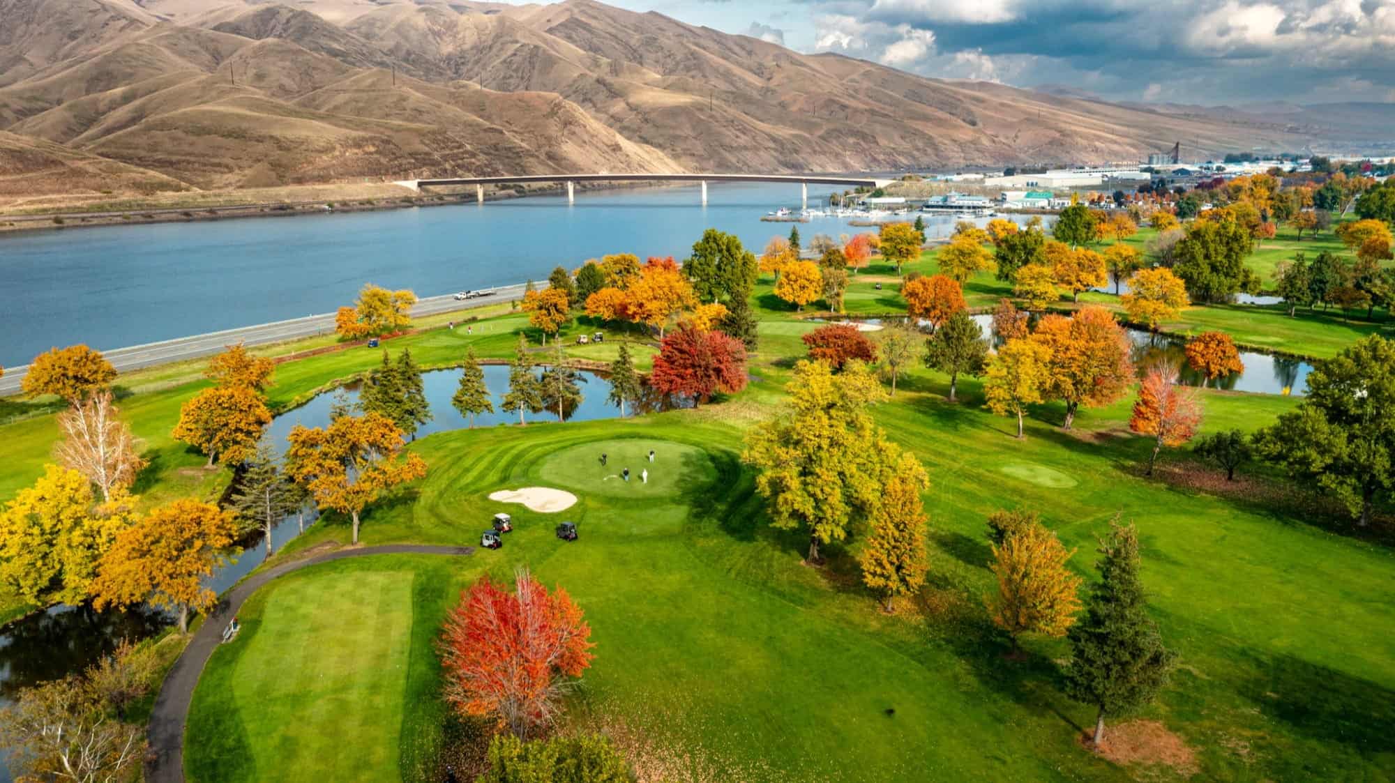 Aerial view of a golf course with autumn trees, a river, a bridge, and hills in the background under a partly cloudy sky—an inviting landscape where adventure and relaxation meet amidst natural beauty.