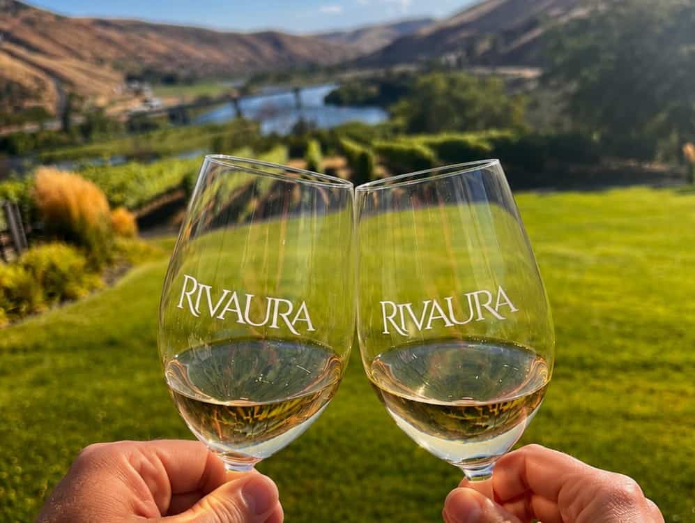 Two hands hold Rivaura wine glasses in a toast, celebrating Northwest wine and culture, with a green lawn, vineyard, river, and hills in the background under a blue sky.