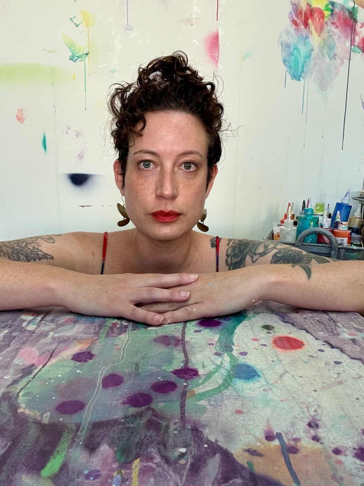 A person with short curly hair and tattoos leans on a colorful, paint-splattered table during Studio Sessions in an art studio filled with various supplies and abstract art on the wall, reminiscent of Liz Tran’s vibrant style.