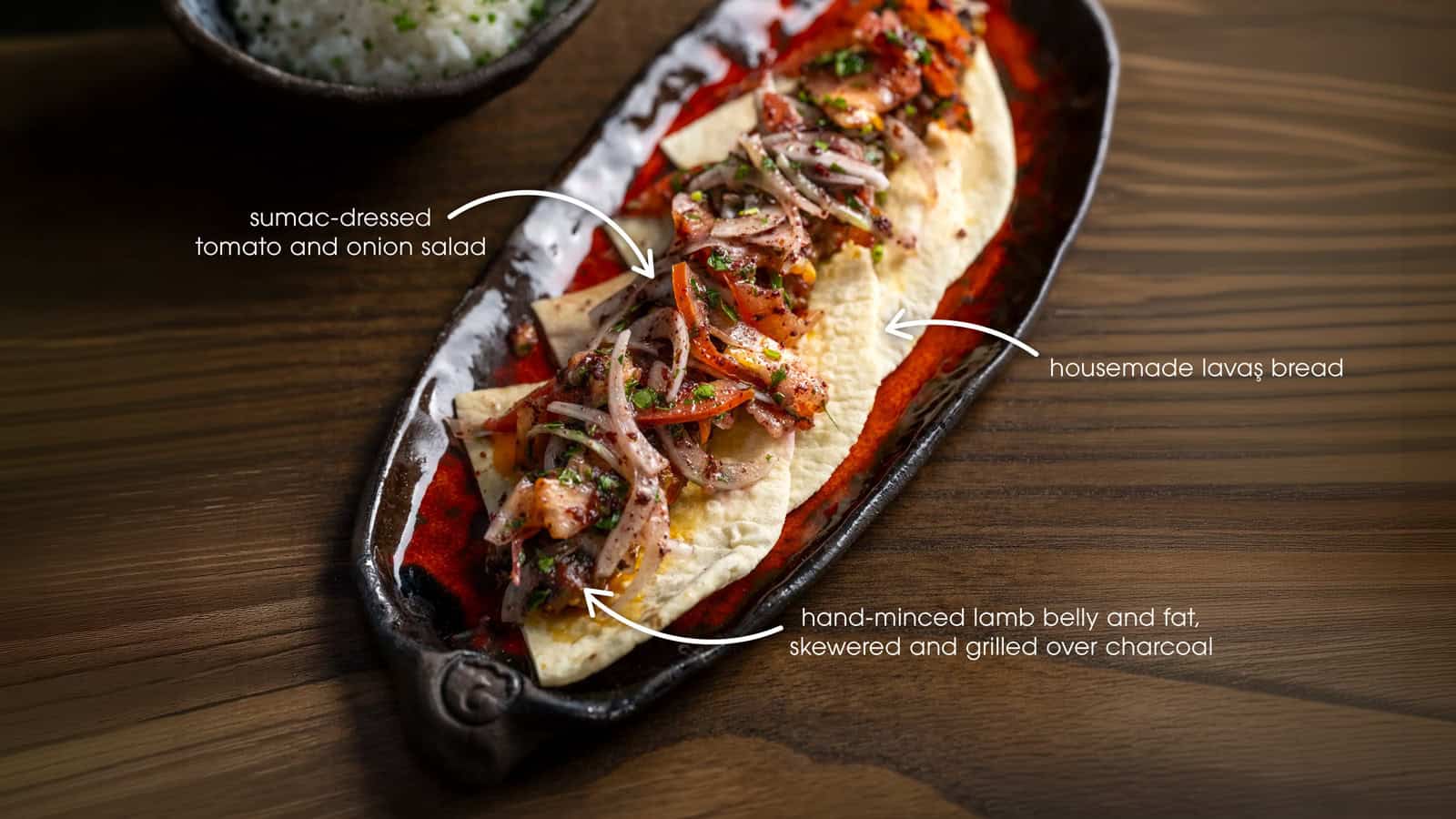 Oval plate with lavash bread topped with grilled minced lamb, tomato and onion salad; dish is labeled with ingredient notes.