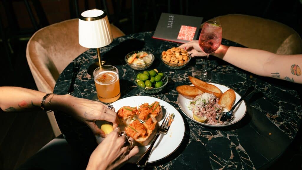 Two people sit at a marble table in a cozy cocktail bar with cocktails, beer, olives, dips, bread, nuts, and pickles under a small lamp, reaching for food.