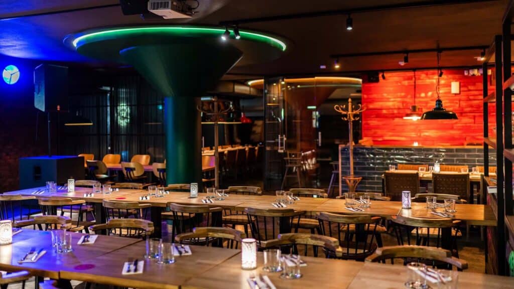 Modern restaurant interior with wooden tables set with glasses and cutlery, warm lighting, and a prominent green column in the center—an inviting space reminiscent of Helsinki’s contemporary dining scene.