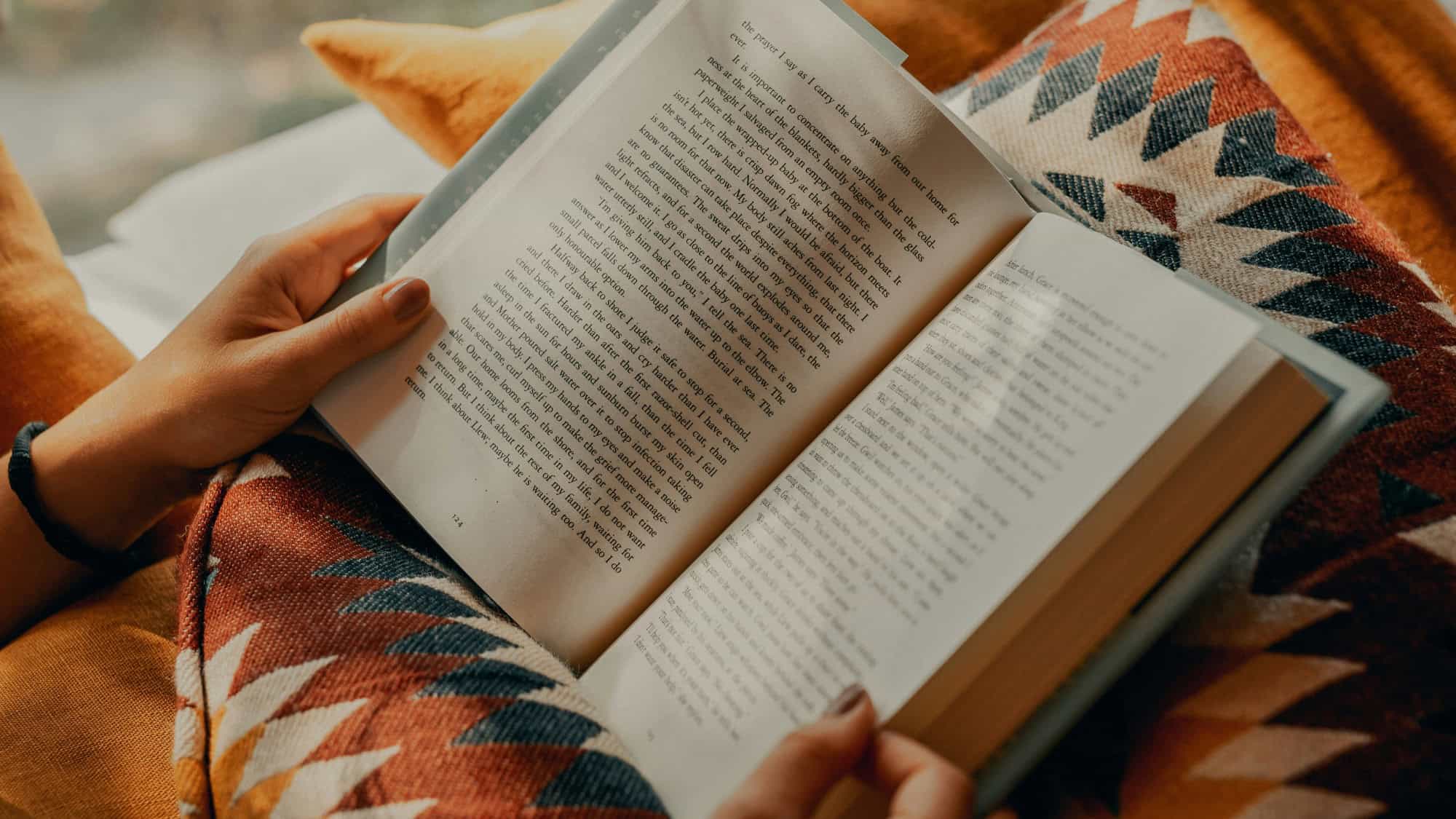 Person holding and reading an open book while sitting under an orange patterned blanket.