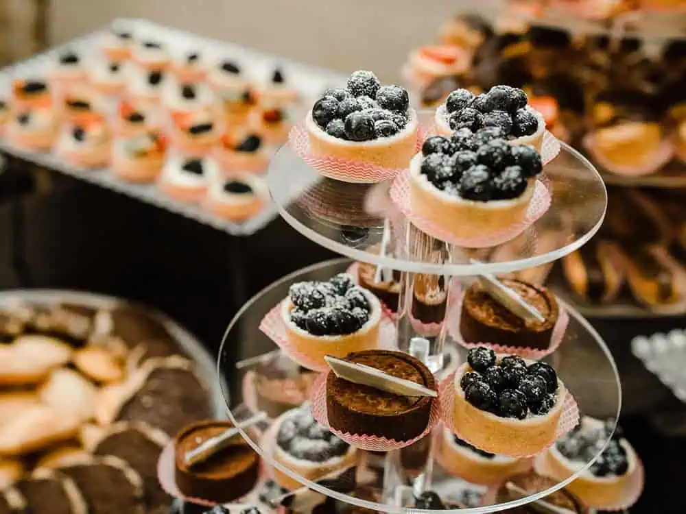 A tiered stand displays assorted mini fruit tarts and chocolate desserts, curated by Herban Feast’s culinary director, with additional pastries blurred in the background.