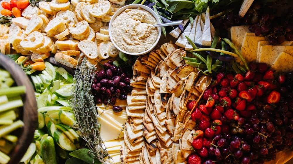 Assorted platter with pita, sliced bread, hummus, cheese, olives, green beans, cucumbers, strawberries, grapes, and tomatoes arranged on a table—perfectly curated with holiday hosting tips inspired by the culinary director at Herban Feast.