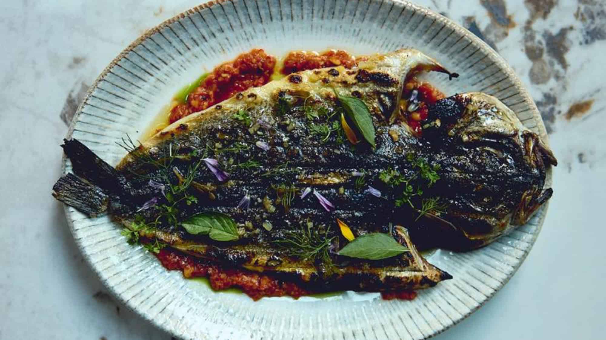 Grilled whole fish served on a ceramic plate, garnished with fresh herbs and placed over a bed of red tomato-based sauce.