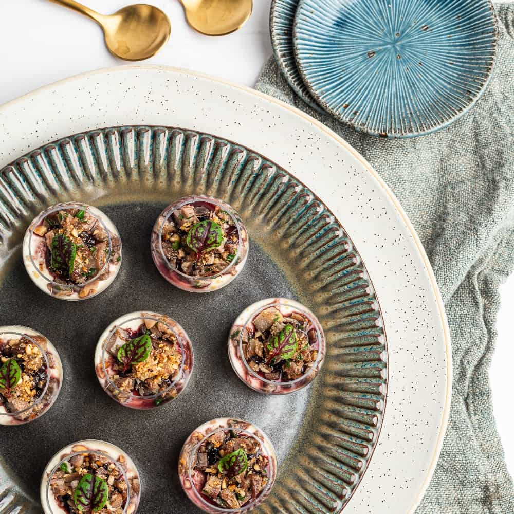 Seven small dessert cups topped with granola and herbs are arranged on a decorative plate beside two gold spoons and blue plates on a textured fabric.