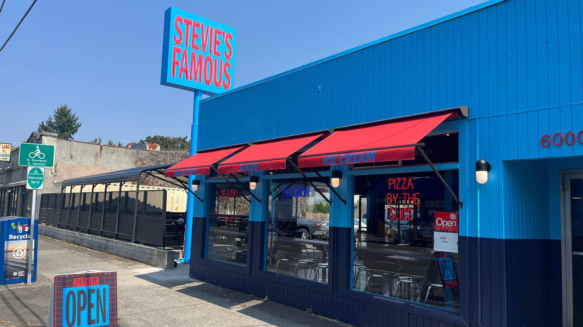 A blue restaurant with red awnings reads “Stevie’s Famous.” Signs advertise ice cream, pizza by the slice, and display “OPEN” near the entrance. Outdoor seating is visible—perfect for enjoying a bite in North Seattle.