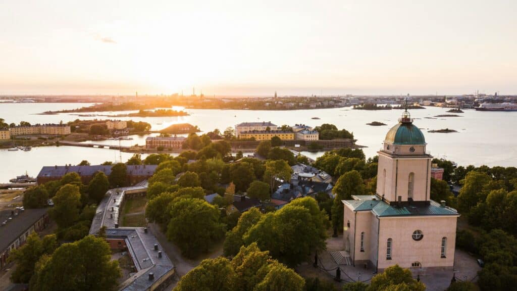 Aerial view of a church with a green dome surrounded by trees, overlooking water and islands at sunset.
