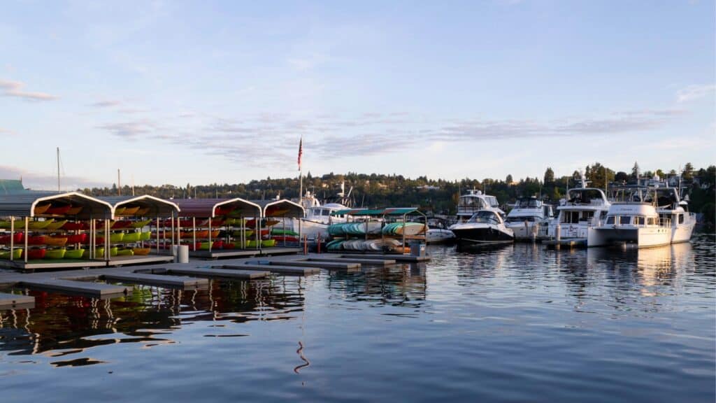 Rows of colorful kayaks under shelters and several yachts docked at a marina, with calm water and trees in the background under a partly cloudy sky.