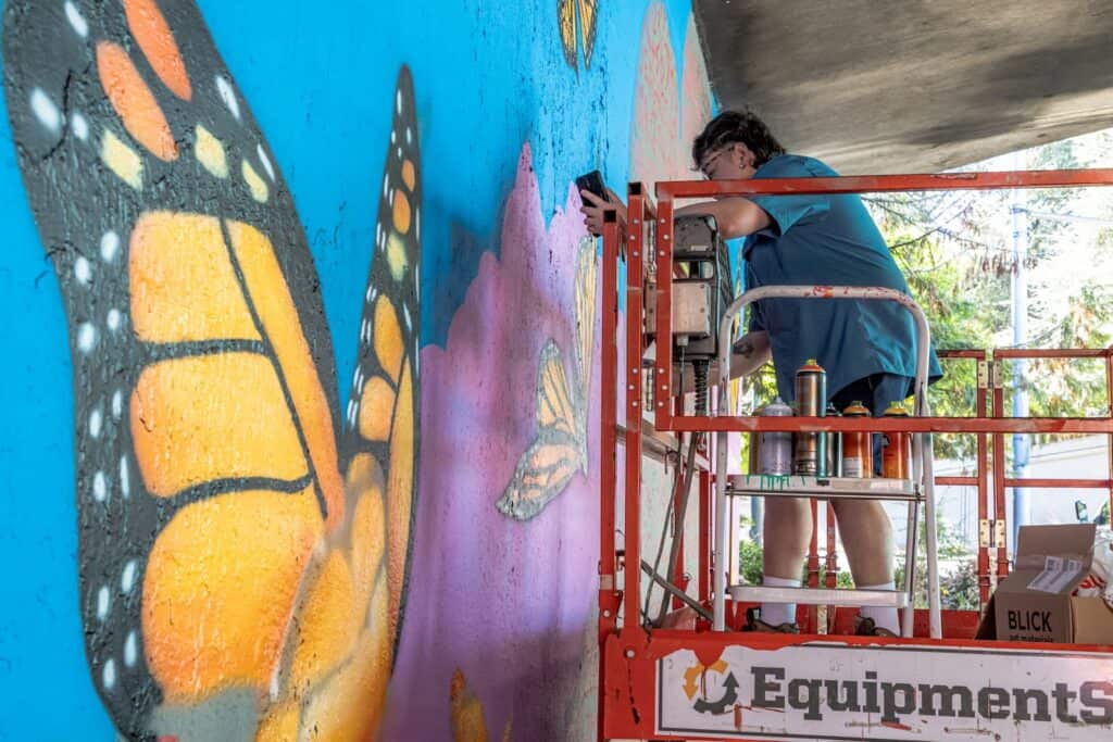 A person on a lift paints a mural of a butterfly on a wall, with spray paint cans and supplies visible nearby.