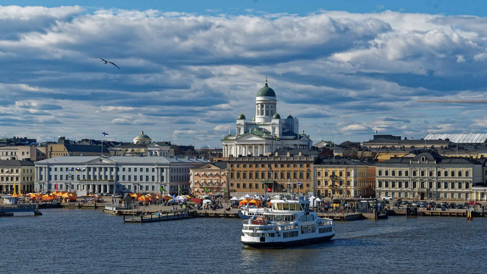 A white ferry crosses the harbor in front of historic buildings and Helsinki Cathedral under a partly cloudy sky.