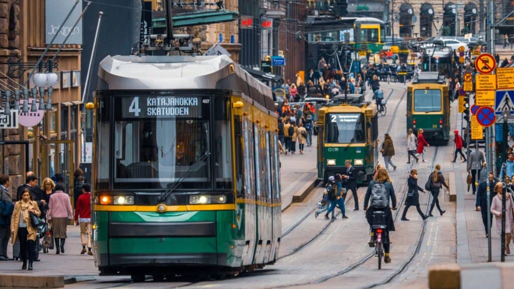A busy city street with trams, cyclists, and pedestrians moving along tracks and sidewalks in an urban center.