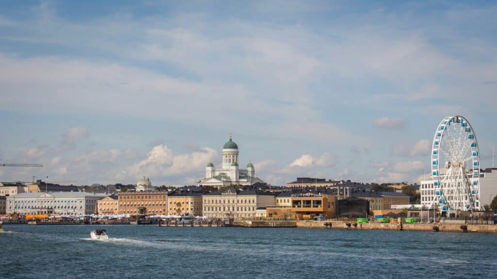 A waterfront cityscape with historic buildings, a large white cathedral with green domes, a Ferris wheel, and a small boat on the water under a partly cloudy sky.
