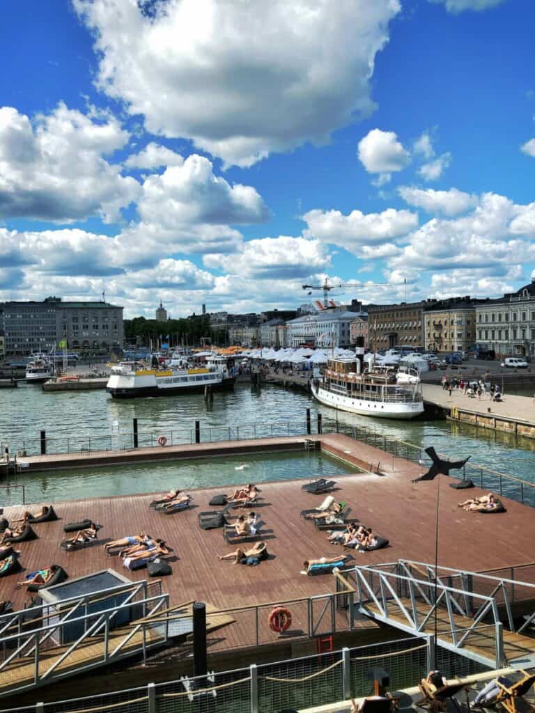 People sunbathe on a wooden deck by the waterfront, with boats docked and historic buildings in the background under a partly cloudy sky.
