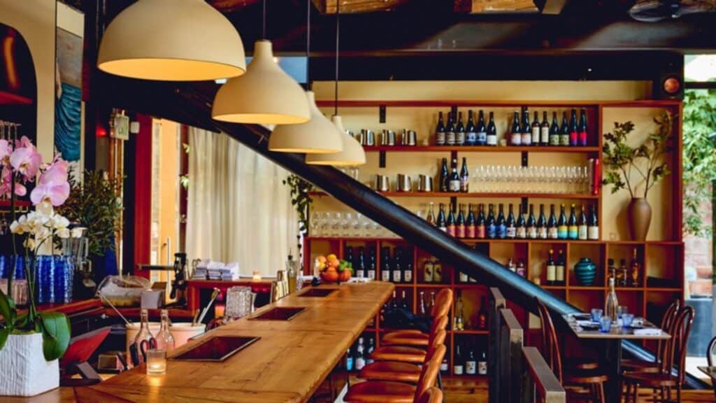 A modern bar with wooden countertops, pendant lights, bar stools, and shelves stocked with bottles and glasses, with plants and natural light.