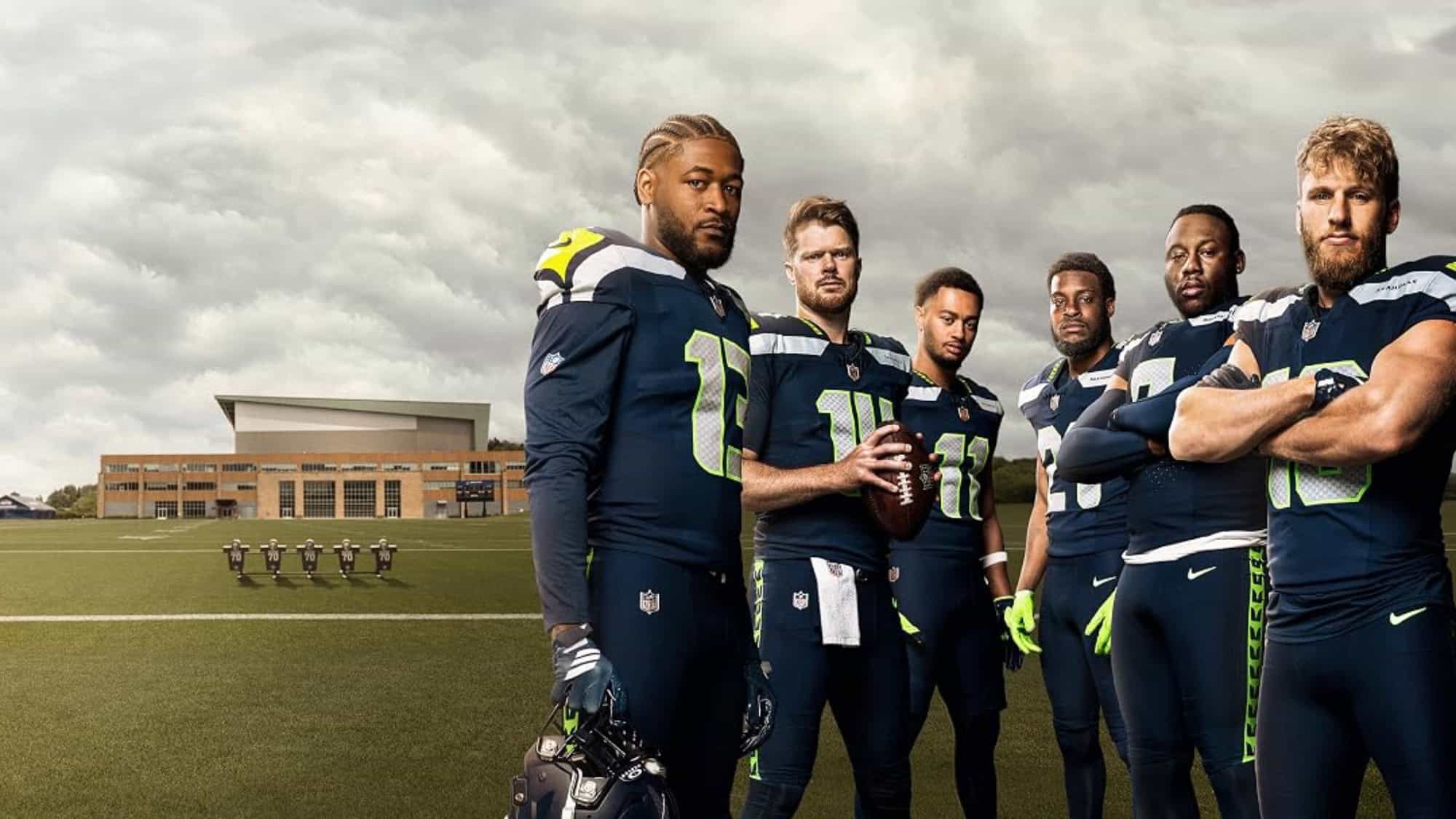 Six American football players in navy and green uniforms, reminiscent of the Seattle Seahawks, stand on a football field with a building in the background under a cloudy sky.