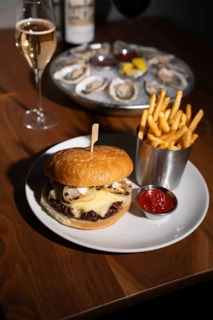A plate with a cheeseburger and fries with ketchup, a glass of white wine, and a tray of oysters on a wooden table.