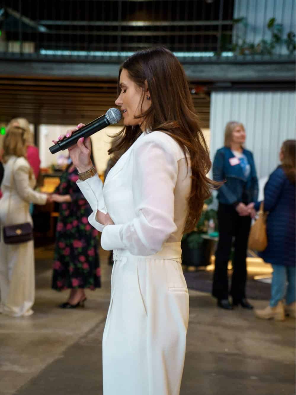 Woman in a white suit speaks into a microphone at an indoor Seattle event. Several people are visible in the background, engaging in conversation.