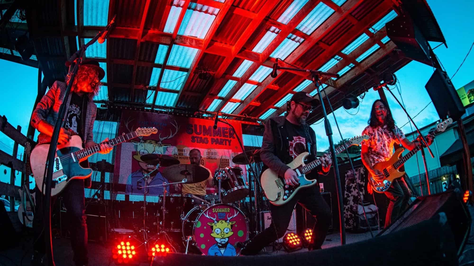 Four musicians play guitar, bass, and drums on an outdoor stage under a red light, with a “Summer Stag Party” banner and cartoon character art in the background.