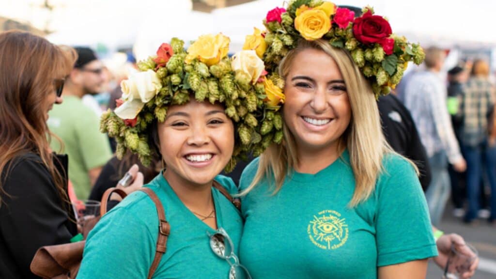 Two women wearing green shirts and floral hop crowns smile at an outdoor event with a crowd visible in the background.