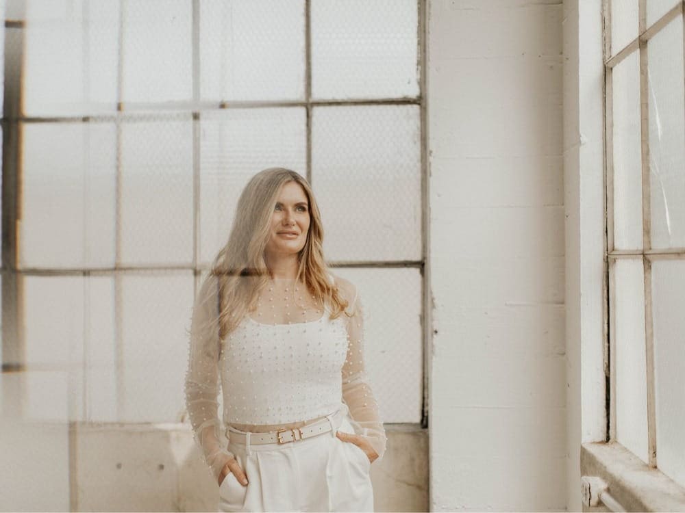 Woman with long blonde hair stands indoors near large windows in Seattle, wearing a sheer long-sleeve top and high-waisted white pants with hands in pockets, changing the conversation through her confident and modern style.