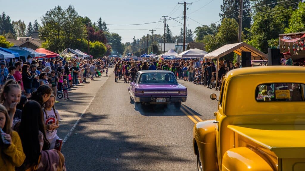 A purple car drives down a crowded street during a parade or car show, with people watching from both sides and tents set up along the road.