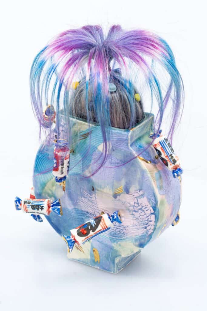 A ceramic vase with a colorful, abstract design, topped with a tuft of blue and purple synthetic hair and decorated with wrapped candies attached to its surface.