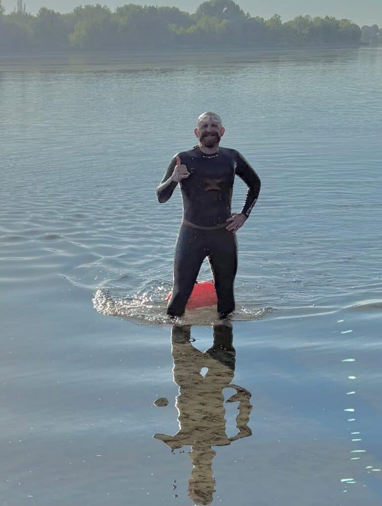 A person in a black wetsuit stands in shallow water, giving a thumbs-up, embodying the spirit of endurance, with trees and a lake visible in the background.