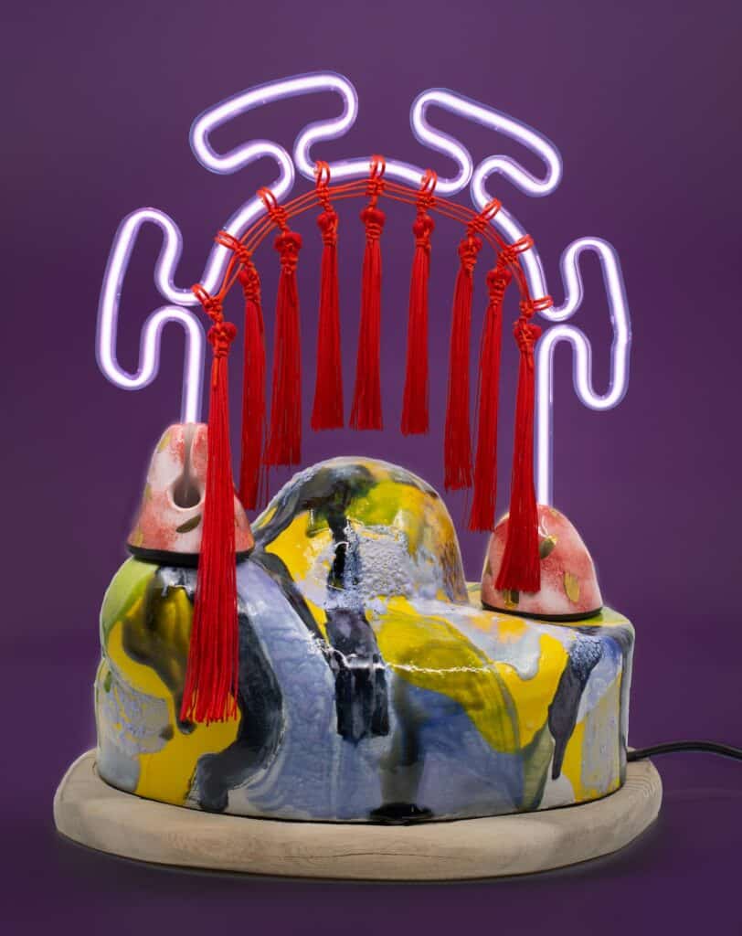 Colorful ceramic sculpture with a wavy surface, topped by a neon arch and hanging red tassels, set against a purple background.