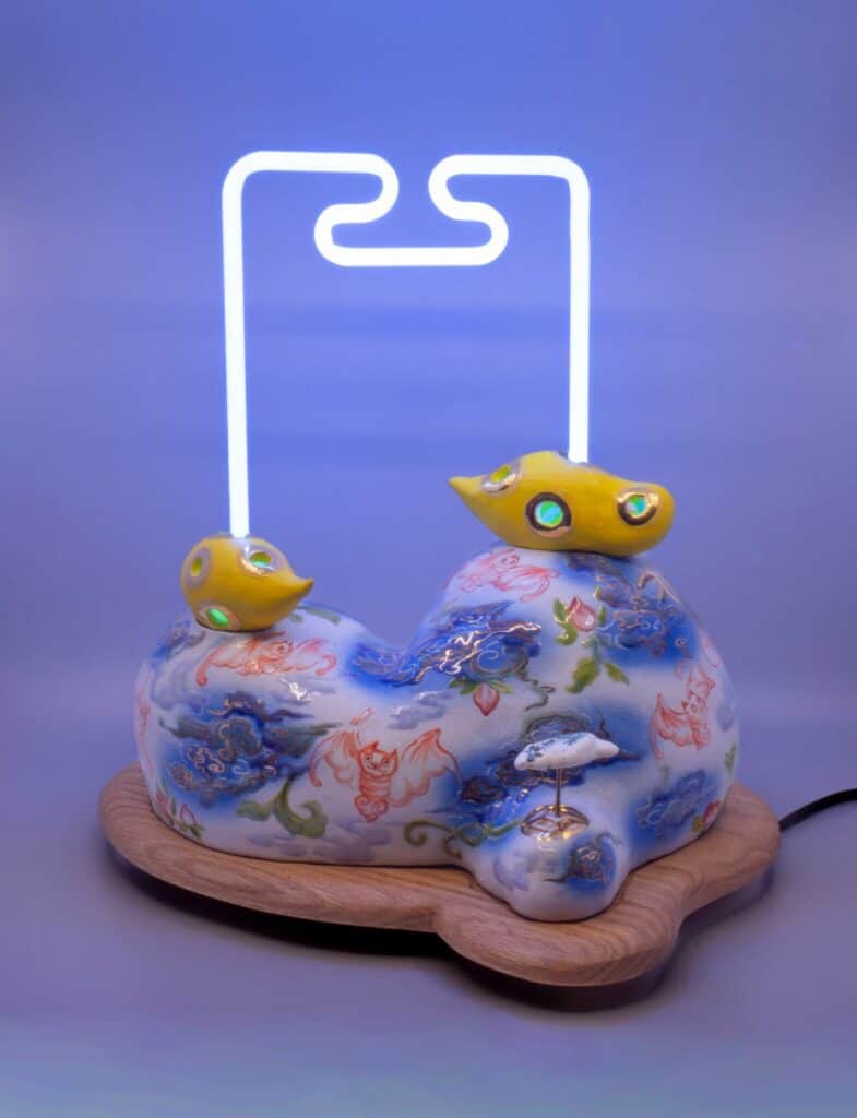 Ceramic base with blue and red patterns, two yellow bird figures, a small crane, and a geometric white neon light on a wooden platform.