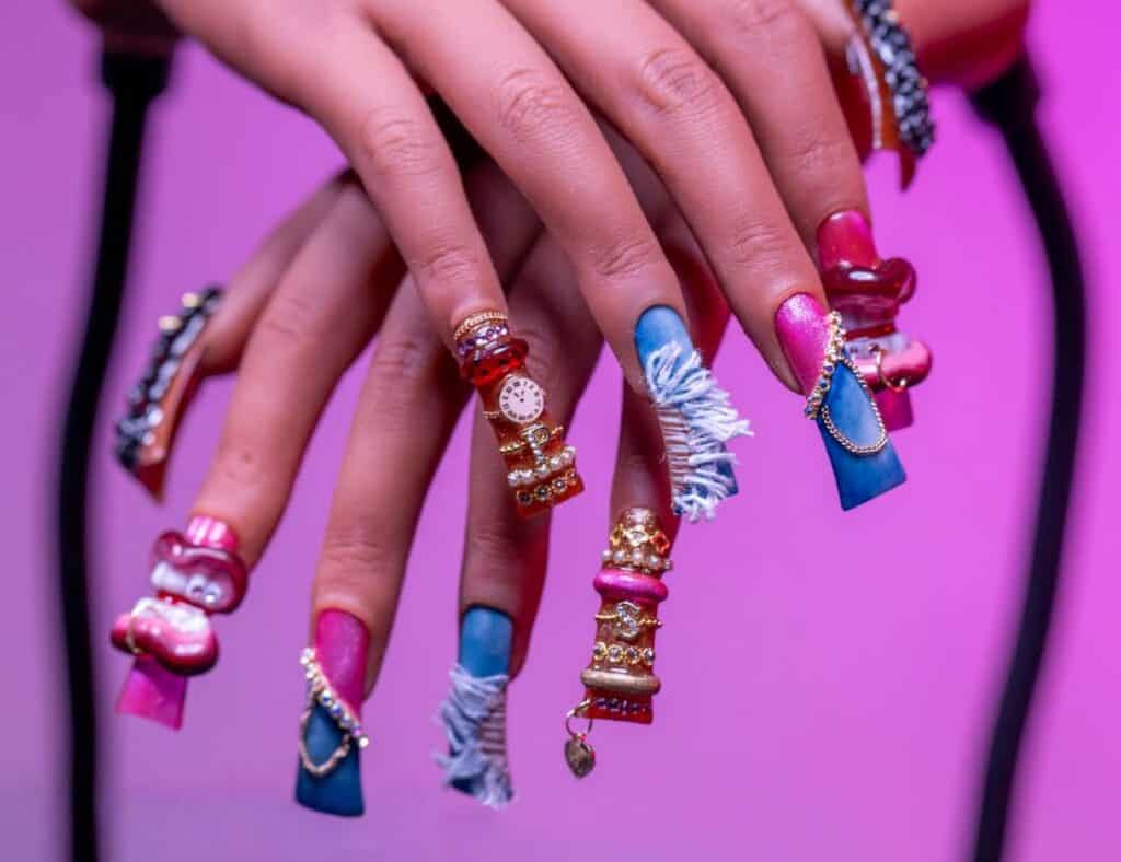 Hands with long, decorated acrylic nails featuring blue and pink polish, 3D embellishments, denim fabric accents, and gold jewelry—channeling the bold aesthetic of Black Women in Hip-Hop Fashion—set against a pink background.