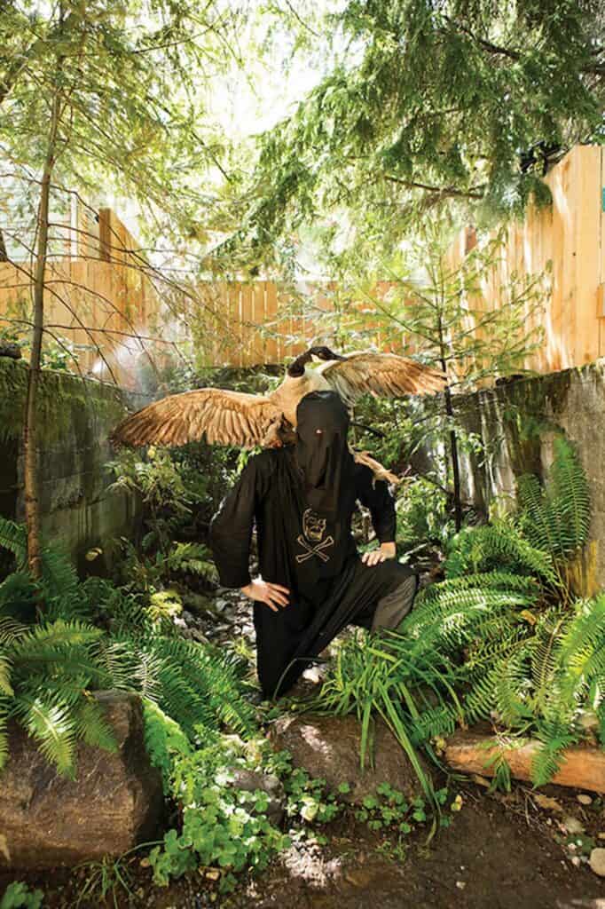 A person in a black hooded robe kneels in a lush, green forest area with a taxidermy goose mounted on their shoulders, evoking an otherworldly scene reminiscent of installations featured at the Frye Art Museum and capturing the spirit of Seattle visual arts 2026.