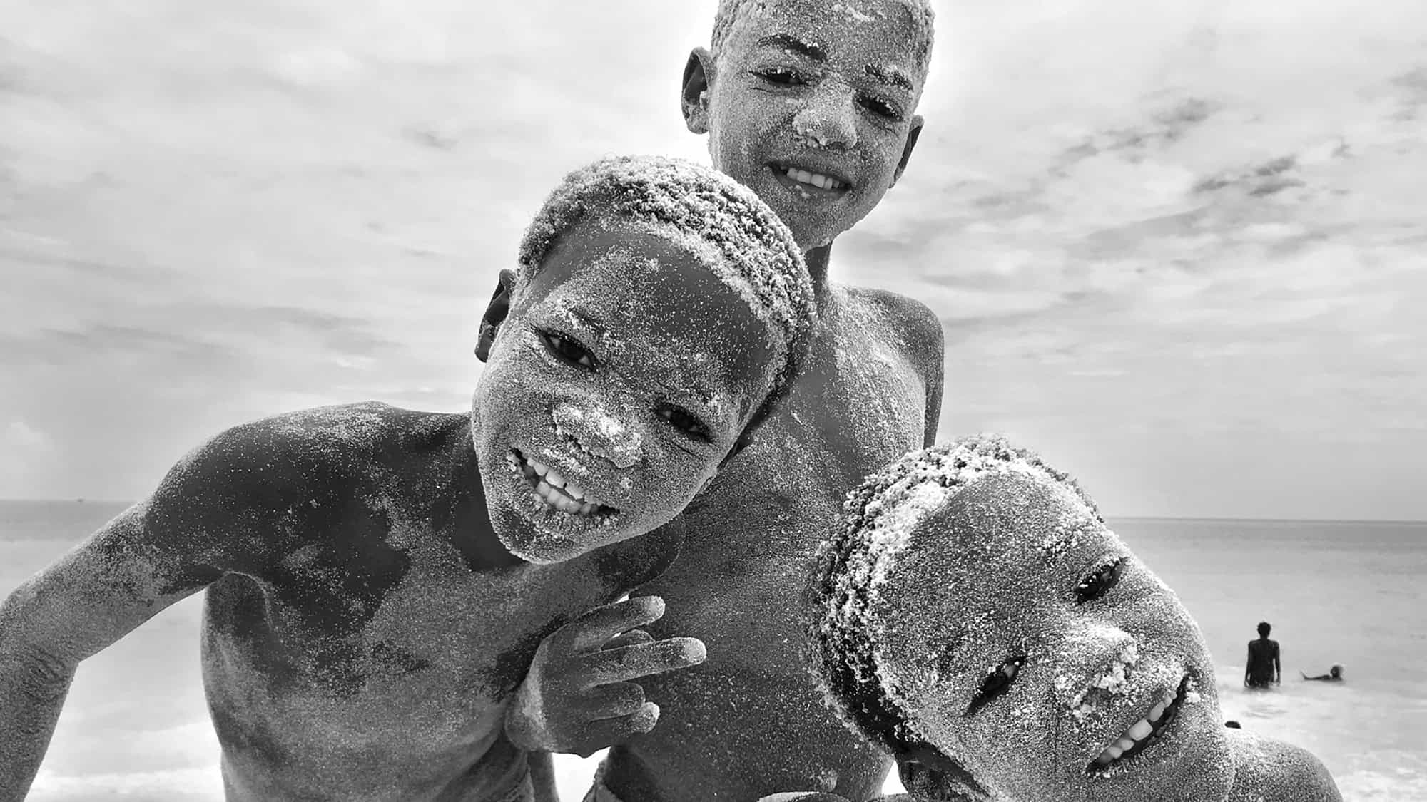 Three children with sand-covered faces and hair smile and pose on a beach, with the ocean and another person visible in the background under a cloudy sky.