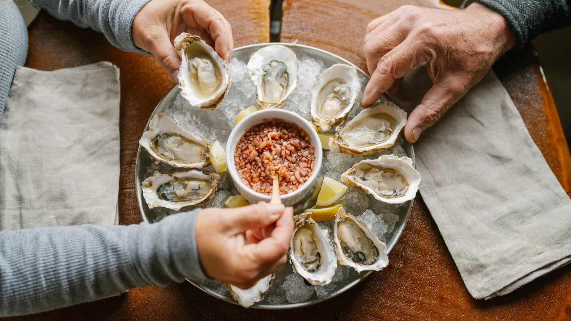 A plate of oysters on ice surrounds a bowl of sauce, with two people using small forks to eat the oysters.