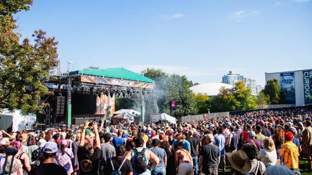A large outdoor crowd faces a stage at the Bumbershoot festival, with trees, a blue sky, and buildings in the background.