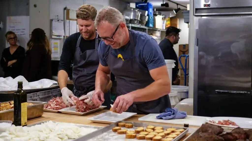 Two men in aprons prepare food in a busy commercial kitchen, slicing meat and arranging ingredients on trays, while others work in the background.