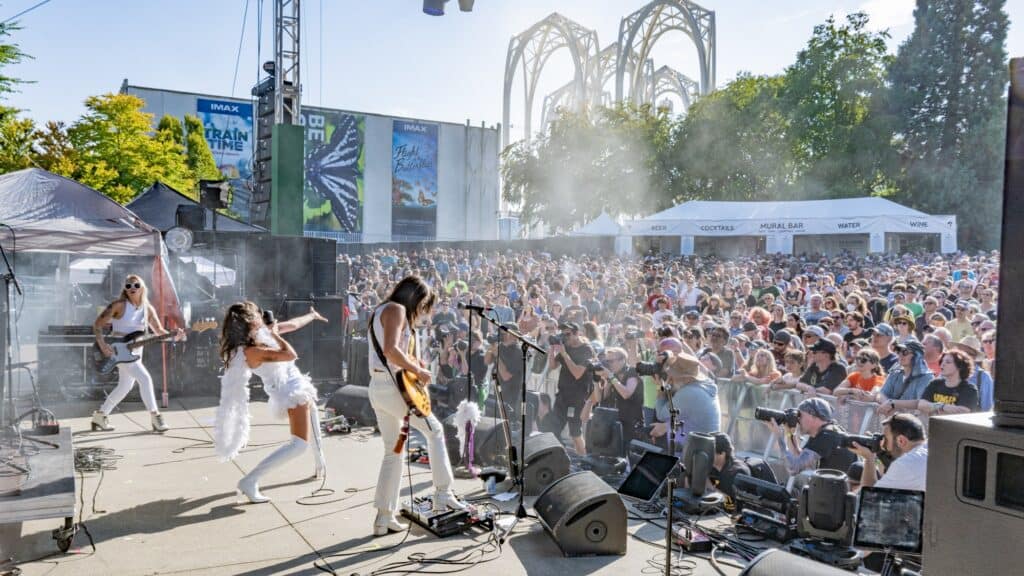 A band performs on an outdoor stage before a large crowd on a sunny day, with trees, tents, and a modern building in the background.