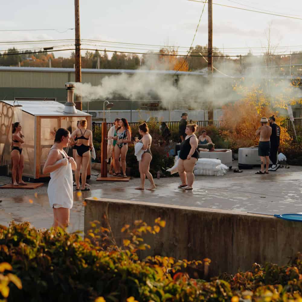 People in swimsuits gather around a steaming outdoor sauna and hot tubs in an urban setting on a sunny day, with some sitting and others standing or walking.