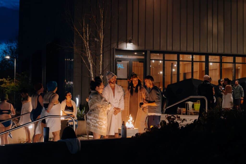A group of people in robes and towels gather around an outdoor fire pit at night outside a modern building with large windows.