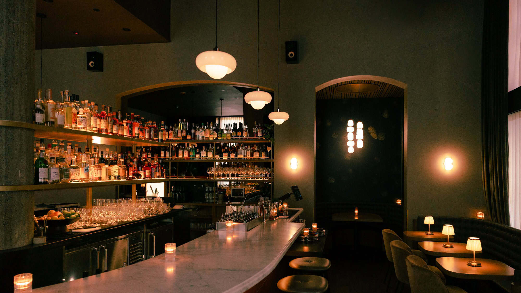 Dimly lit bar with a marble counter, shelves of bottles, pendant lights, and small tables with lamps and chairs.