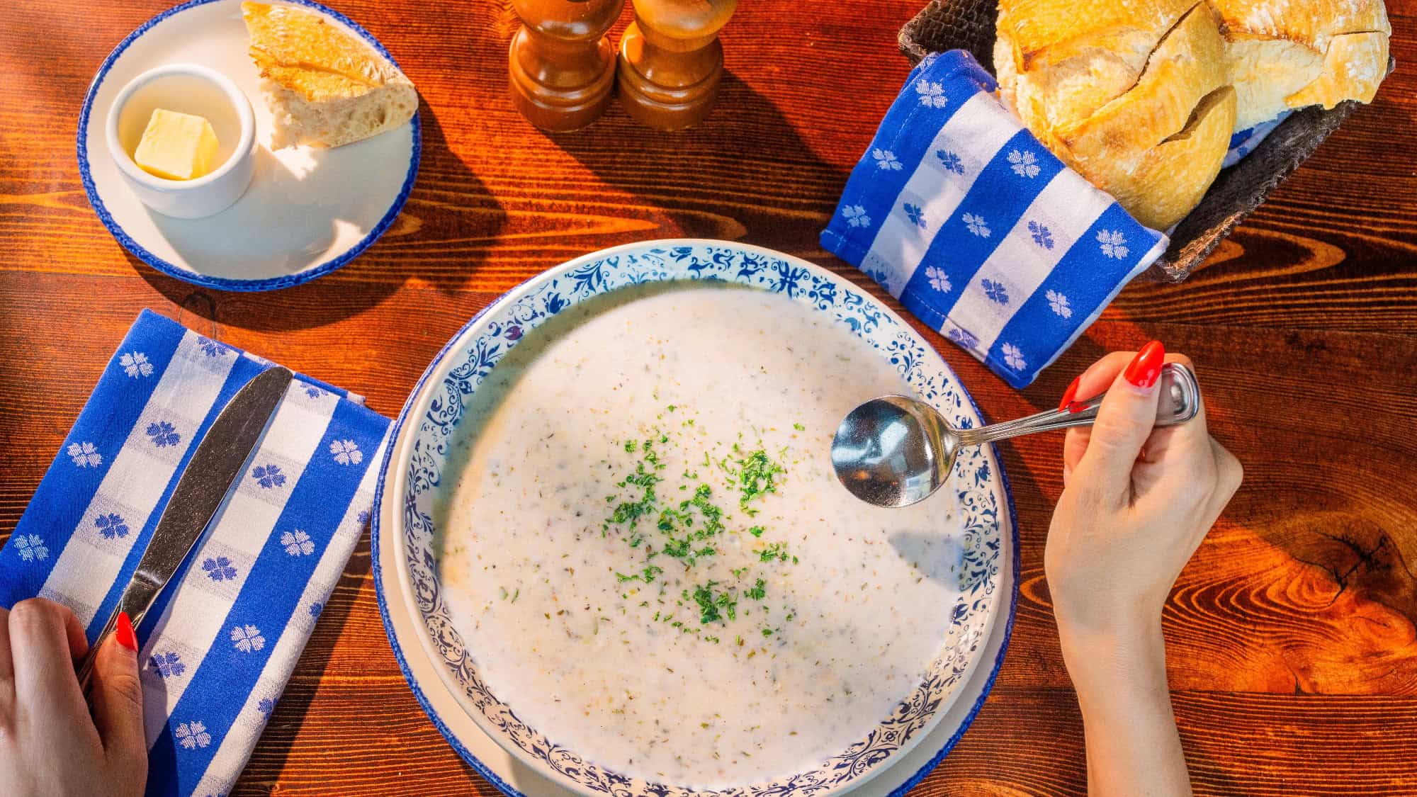 A person holding a spoon over a bowl of creamy soup garnished with herbs, with bread, butter, and blue napkins on a wooden table.