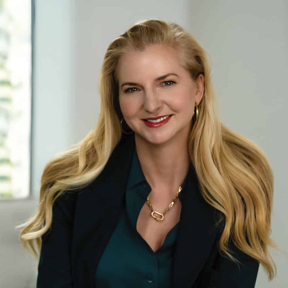 A woman with long blonde hair, wearing a dark blazer, teal blouse, and gold necklace, sits indoors near a window, her smile leaving everyone Blown Away.