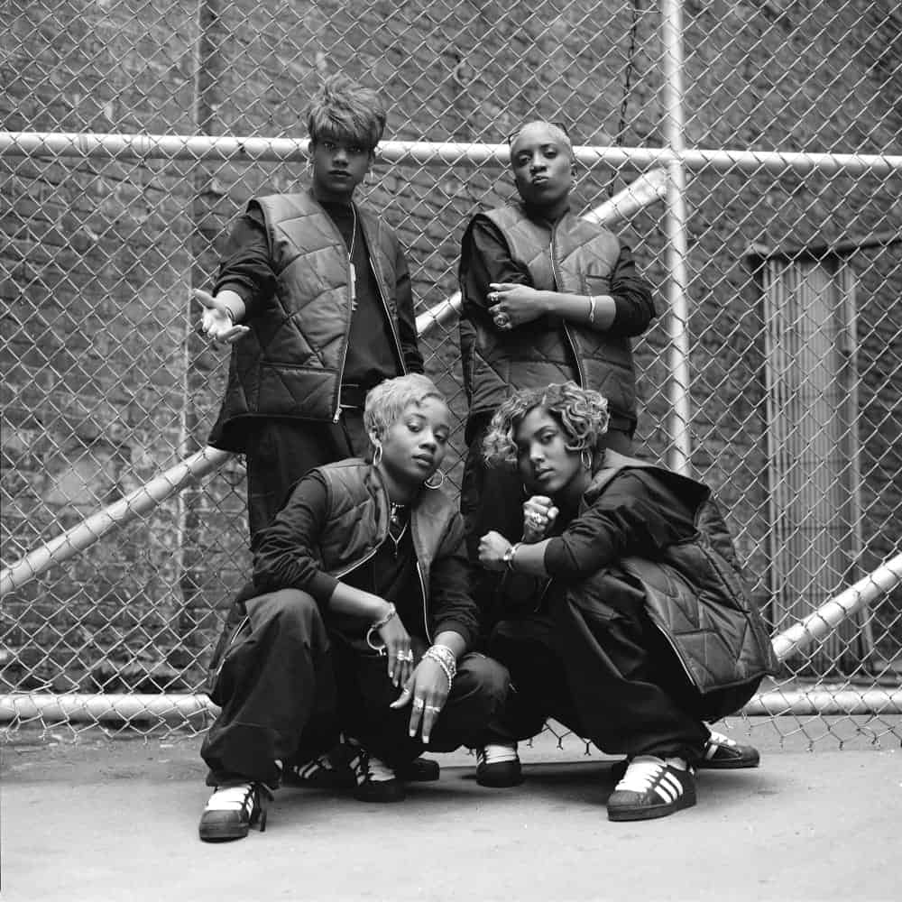 Four people in matching black outfits and vests pose in front of a chain-link fence on a city street, two standing and two crouching—a powerful scene reminiscent of Black Women in Hip-Hop Fashion featured at MoPOP Seattle.