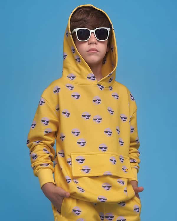 Boy wearing a yellow hoodie with sunglasses emoji print, matching pants, and white sunglasses, standing against a blue background—his look will leave you absolutely blown away.