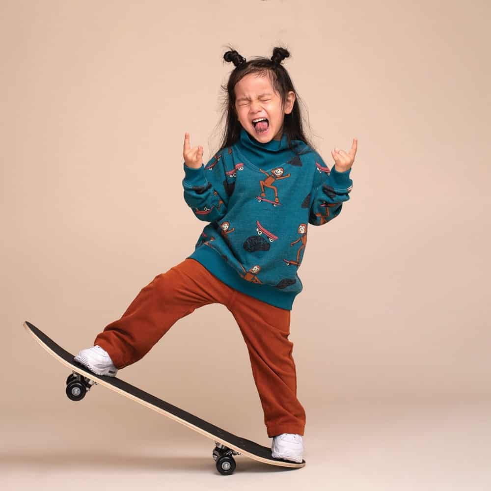 Young child in a blue sweater and brown pants stands on a skateboard, making a playful face with hands raised in a “rock on” gesture, looking absolutely blown away with excitement against a plain beige background.