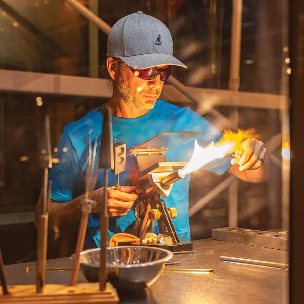 A man wearing a blue shirt and cap uses a torch to shape glass in a workshop, surrounded by tools and rods—a scene reminiscent of moments from Blown Away.