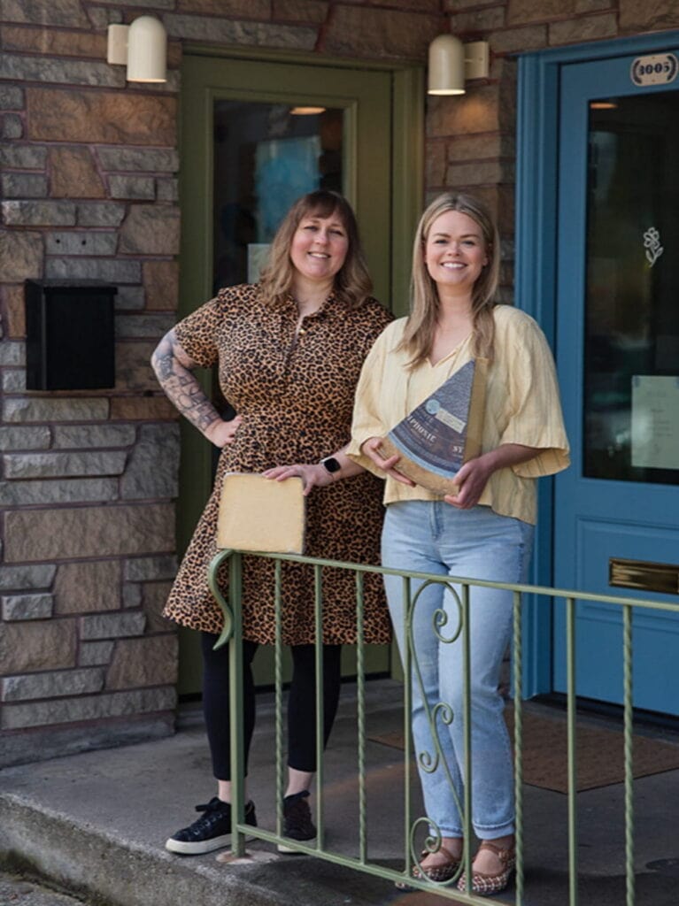 Two women stand in front of a building; one wears a leopard print dress, and the other holds a triangular award. Both are smiling and pose by a green railing, perhaps celebrating their achievements in the vibrant world of Washington artisan cheese.